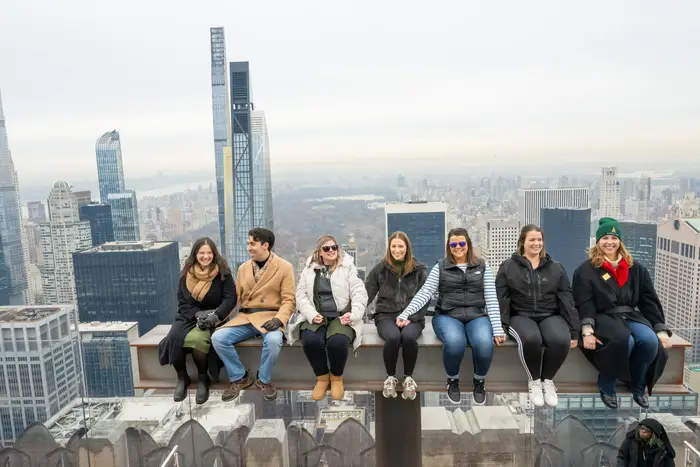 a group of people sit on a beam with a view of the New York skyline behind it.
